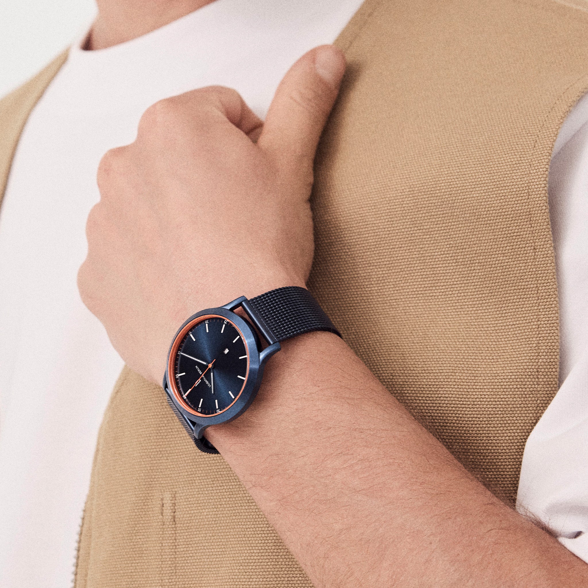 Stylish blue wristwatch with mesh strap and orange bezel on man's wrist in beige vest