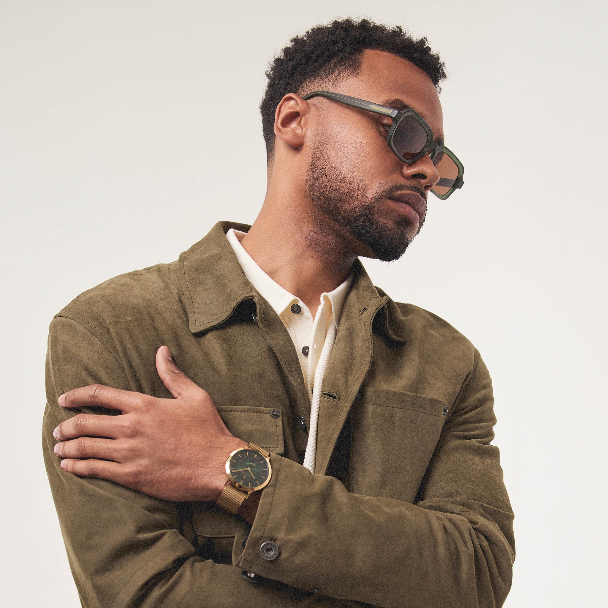 Man in olive jacket wearing square sunglasses and gold wristwatch against neutral background