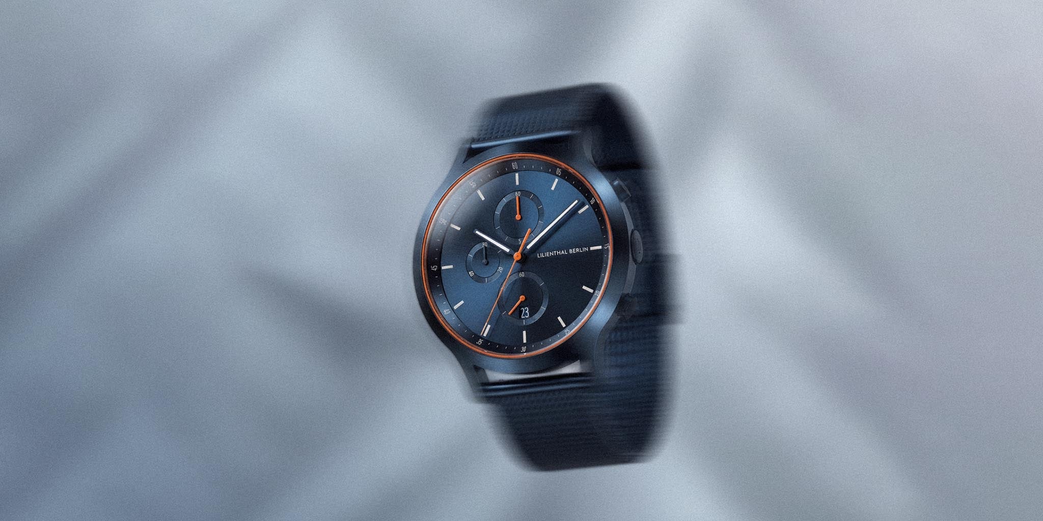 Blue and orange chronograph watch with mesh strap and minimalist dial on gray background