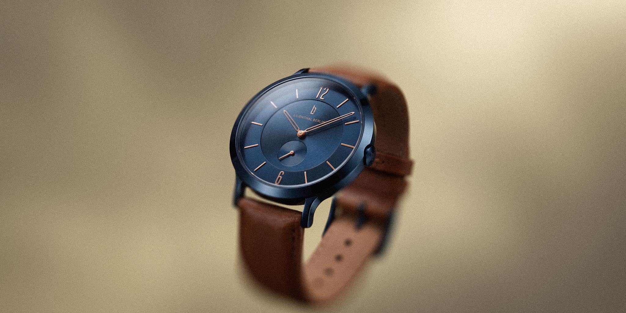 Minimalist blue watch with rose gold accents and brown leather strap on neutral background