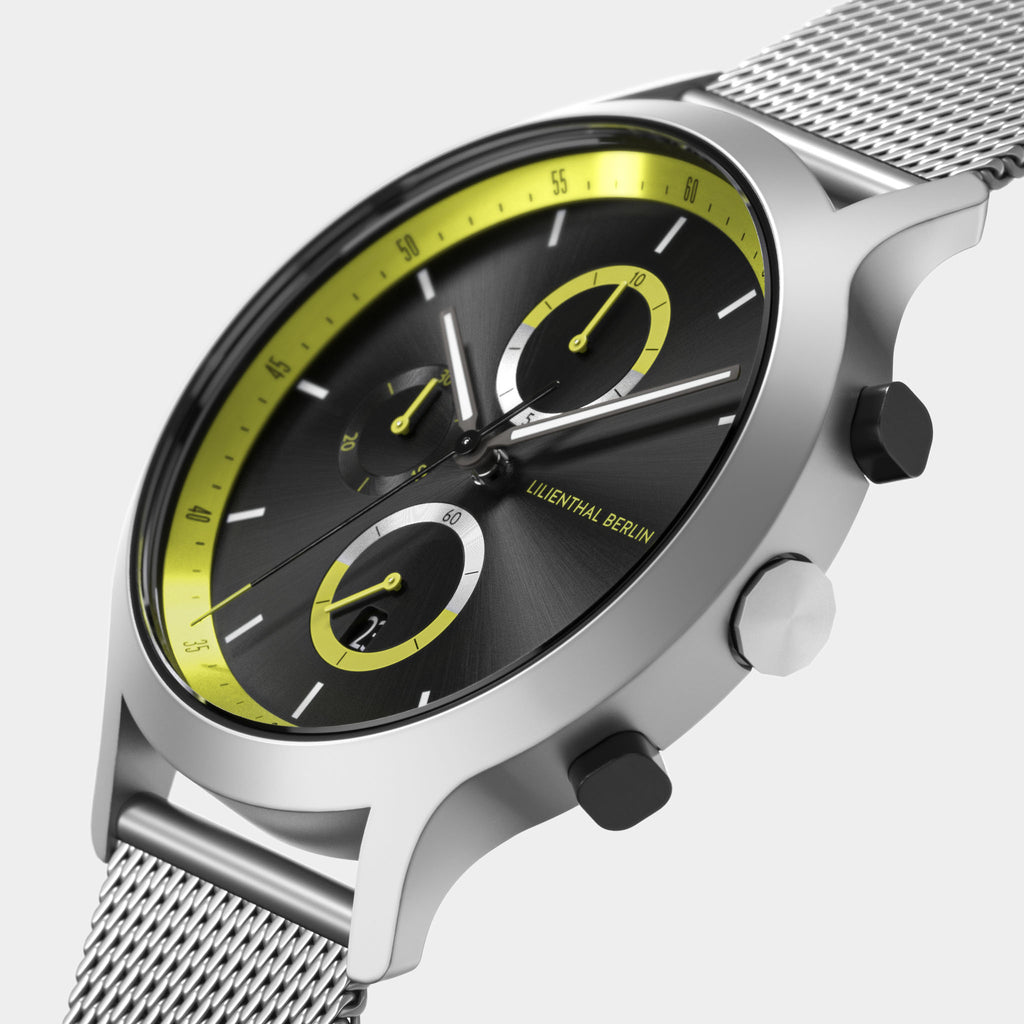 Modern silver mesh watch with black dial, yellow accents, and chronograph subdials