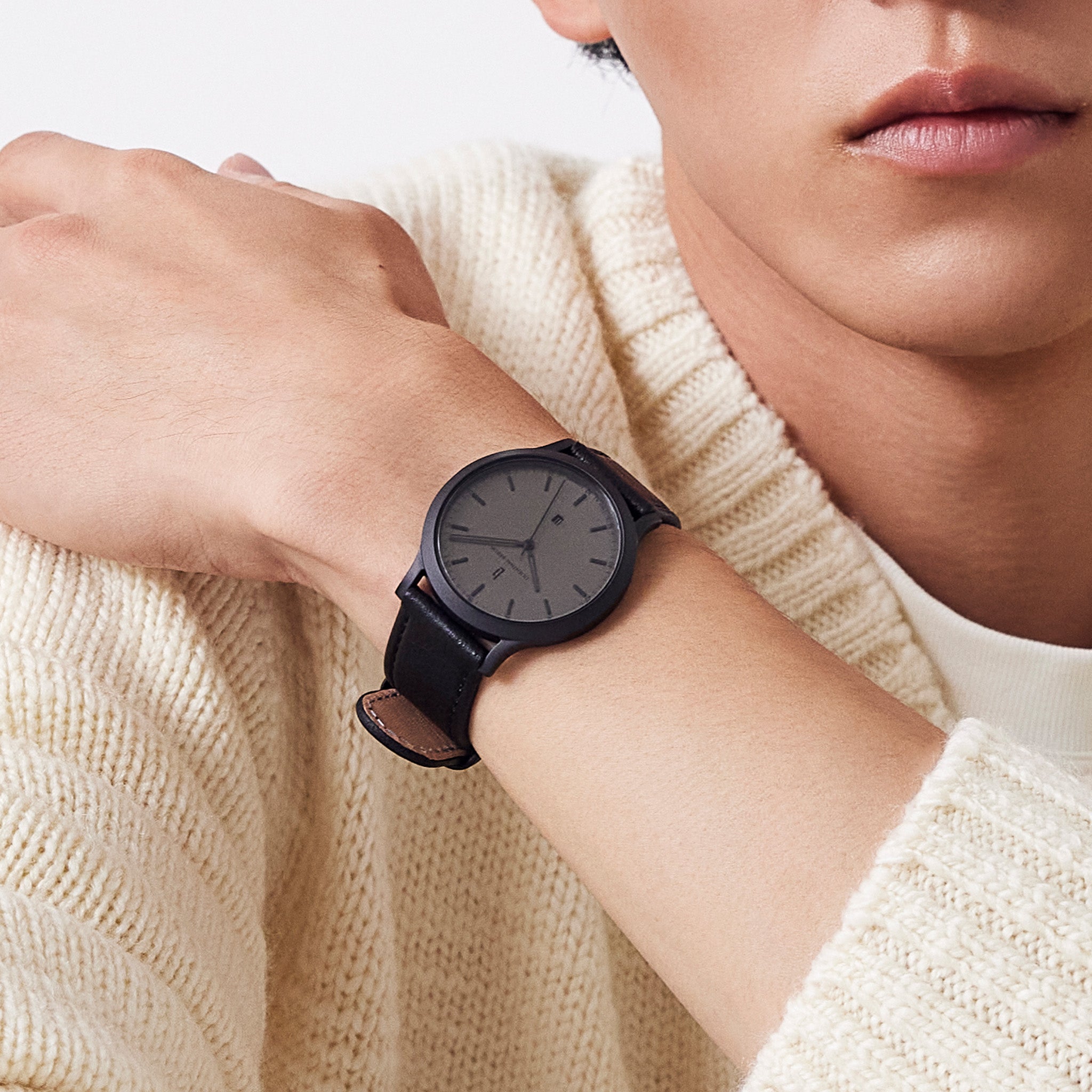Minimalist black wristwatch with leather strap on wrist, worn with cream knit sweater