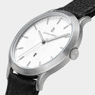 Minimalist silver wristwatch with white dial, black leather strap, and date display