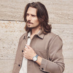 Man in beige jacket wearing modern wristwatch with blue strap, standing by stone wall